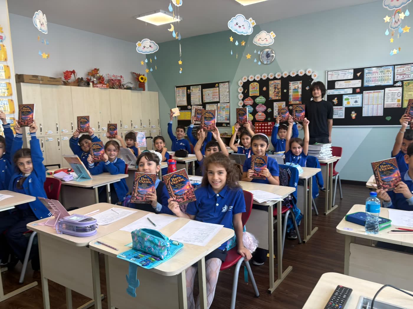 A full classroom of pupils in blue school uniforms holding up copies of the book, with Jamal standing at the back by the board.