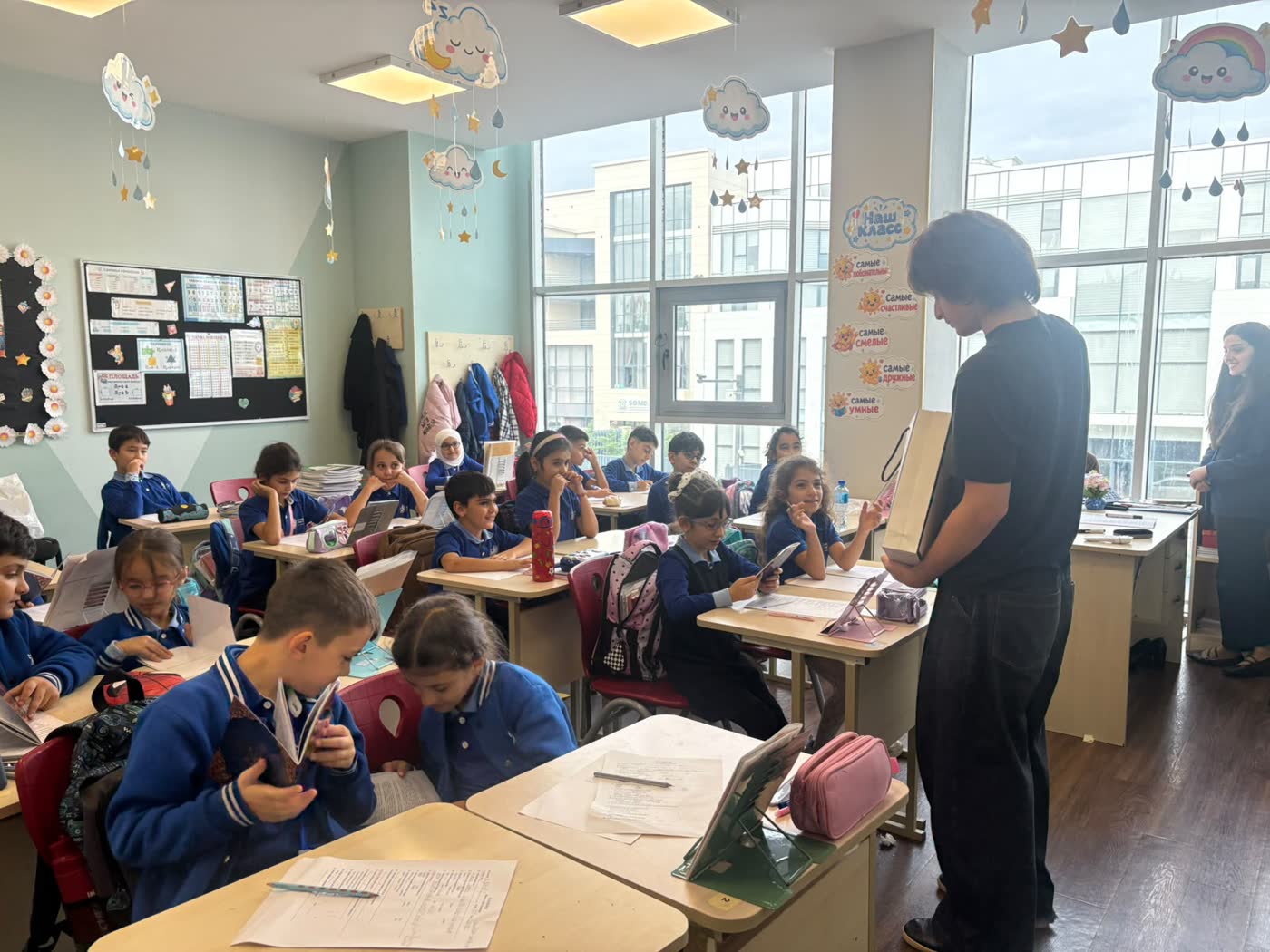 Jamal, seen from behind, holds a bag of books and talks to a class seated at their desks.